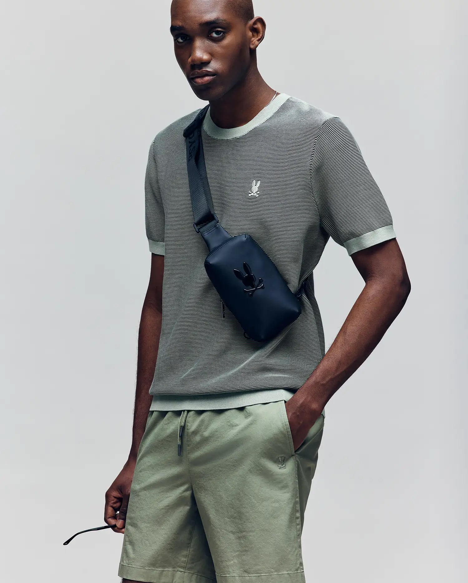 A man wears a sage green Psycho Bunny MENS MARIO STRIPED SWEATER T-SHIRT (MT0400009) with matching shorts and a black crossbody bag featuring an embroidered bunny logo, standing against a plain light gray background.
