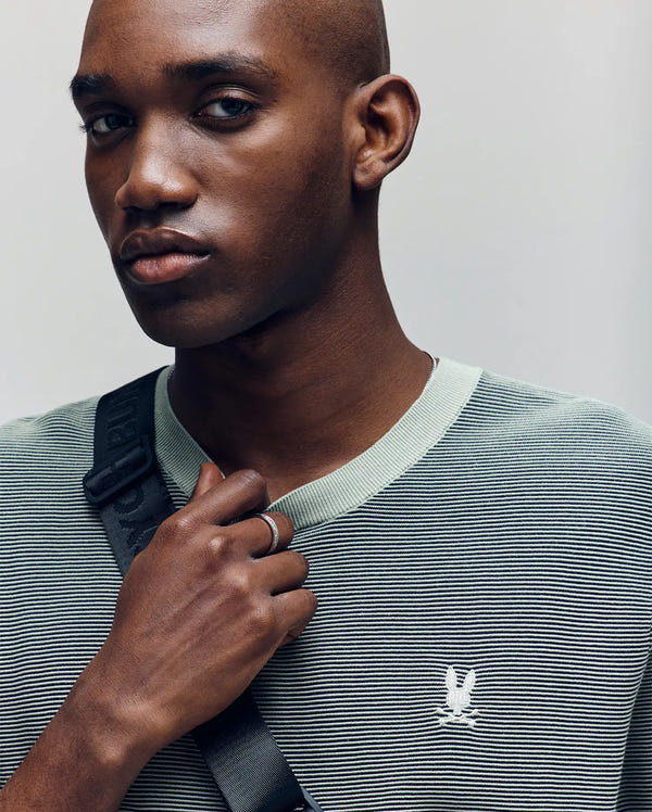 A man with a shaved head wears the Psycho Bunny MENS MARIO STRIPED SWEATER T-SHIRT - MT0400009 in light green, featuring textured stripes and an embroidered bird logo. He holds a black strap over his shoulder and gazes to the side with a neutral expression.