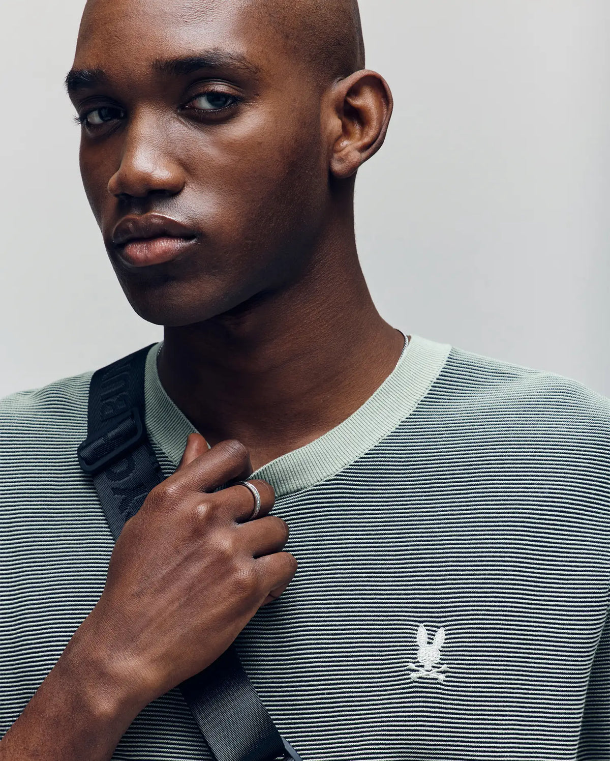 A man with a shaved head wears the Psycho Bunny MENS MARIO STRIPED SWEATER T-SHIRT - MT0400009 in light green, featuring textured stripes and an embroidered bird logo. He holds a black strap over his shoulder and gazes to the side with a neutral expression.
