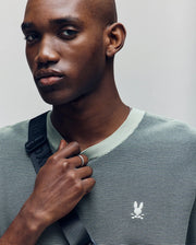 A man with a shaved head wears the Psycho Bunny MENS MARIO STRIPED SWEATER T-SHIRT - MT0400009 in light green, featuring textured stripes and an embroidered bird logo. He holds a black strap over his shoulder and gazes to the side with a neutral expression.