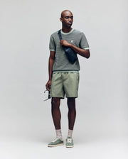 A man in a Psycho Bunny MENS MARIO STRIPED SWEATER T-SHIRT (MT0400009), light green shorts, green sneakers, and light socks stands holding sunglasses and wearing a black crossbody bag against a plain light background.