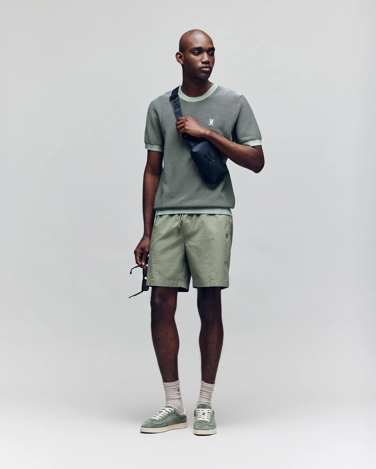 A man in a Psycho Bunny MENS MARIO STRIPED SWEATER T-SHIRT (MT0400009), light green shorts, green sneakers, and light socks stands holding sunglasses and wearing a black crossbody bag against a plain light background.