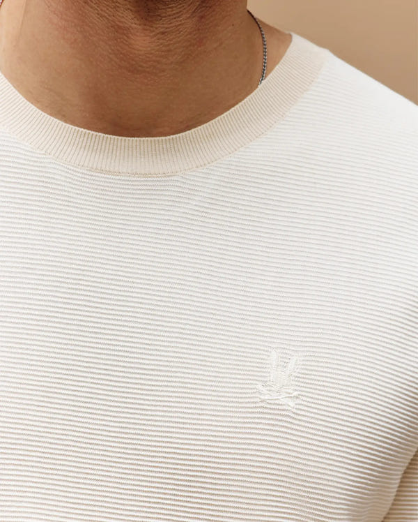 Close-up of a person in the Psycho Bunny MENS MARIO STRIPED SWEATER T-SHIRT (MT0400009), featuring cream-colored ribbed knit with a small embroidered logo on the left chest for a subtle retro touch; only lower face, neck, and upper chest are shown.