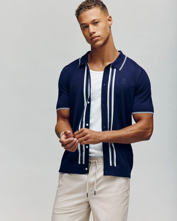 A man with short curly hair exudes retro charm in the Psycho Bunny MENS PATRICE BORDER STRIPE KNIT SHIRT (MT0400054), a navy intarsia-knit short-sleeve button-up with white vertical stripes, worn over a white tee and light pants.