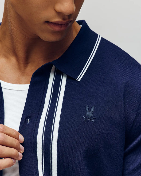 A person wears the Psycho Bunny MENS PATRICE BORDER STRIPE KNIT SHIRT (MT0400054), a navy blue collared shirt with white stripes and rabbit and crossbones logo, crafted from soft knit cotton, layered over a white undershirt; face partly visible.
