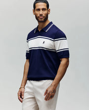 A man with short dark hair and a beard wears the Psycho Bunny MENS ZAVIAN STRIPE KNIT POLO - MT0400055 in navy blue and white horizontal cotton stripes, paired with white shorts, standing against a neutral background.