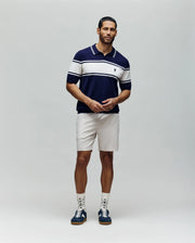 A man with short dark hair and a neat beard wears the Psycho Bunny MENS ZAVIAN STRIPE KNIT POLO - MT0400055, featuring navy and white stripes, paired with light shorts, patterned socks, and navy sneakers against a plain background.
