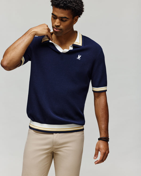 A man wears the Psycho Bunny MENS CARTER SWEATER POLO (MT0400093), a navy cotton short-sleeve polo with a Johnny collar, white and yellow trim, and a white leaf logo, paired with light beige pants against a plain light background.