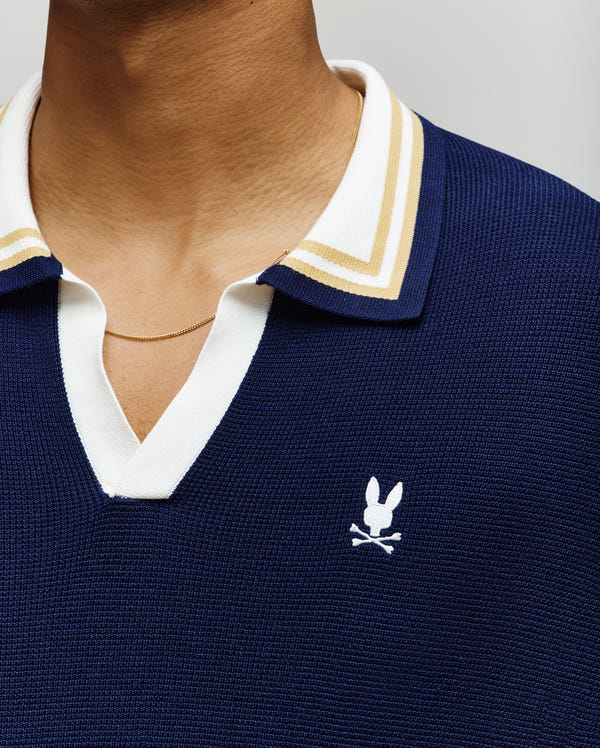 Close-up of a person wearing the Psycho Bunny MENS CARTER SWEATER POLO (MT0400093), a navy cotton knit with a Johnny collar, white and beige striped trim, gold chain necklace, and white embroidered bunny skull and crossbones logo on the chest.