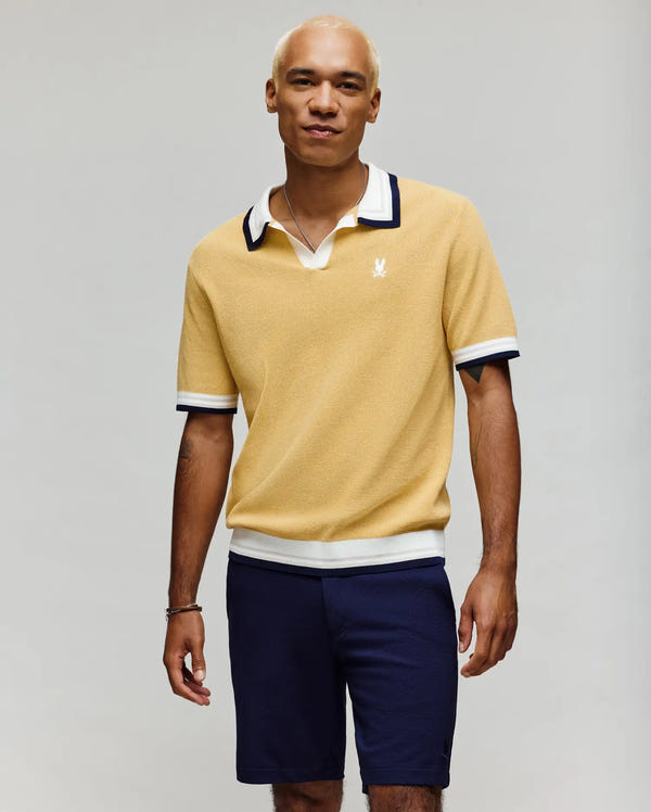 A man with short blond hair wears the Psycho Bunny MENS CARTER SWEATER POLO - MT0400093, a colorblocked yellow short-sleeve cotton knit shirt with a white Johnny collar and navy trim, paired with navy shorts. He smiles at the camera, standing against a light gray background.