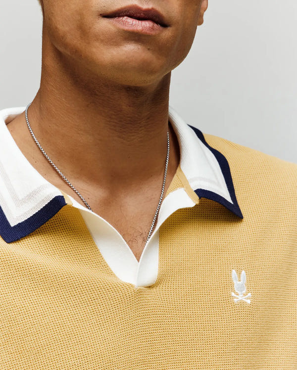 Close-up of a person wearing the Psycho Bunny MENS CARTER SWEATER POLO (MT0400093), a yellow colorblocked Johnny collar shirt with white and navy trim and an embroidered white bunny logo, paired with a thin silver chain necklace. Only the lower half of the face is visible.