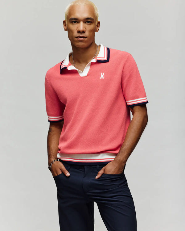 A man with short blond hair wears the Psycho Bunny MENS CARTER SWEATER POLO - MT0400093 in pink, featuring a Johnny collar and white/navy trim, paired with navy pants and a bracelet, standing against a plain light background.