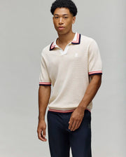 A young man with short, curly hair wears the Psycho Bunny MENS CARTER SWEATER POLO - MT0400093, a light beige regular fit top with a Johnny collar and red, black, and white trim. He pairs it with dark trousers against a light grey background.