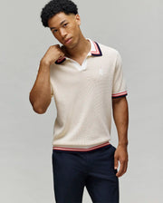 A man poses against a plain background wearing the Psycho Bunny MENS CARTER SWEATER POLO (MT0400093), a regular fit beige short-sleeve polo featuring a Johnny collar and red, navy, white striped cuffs, paired with dark pants.