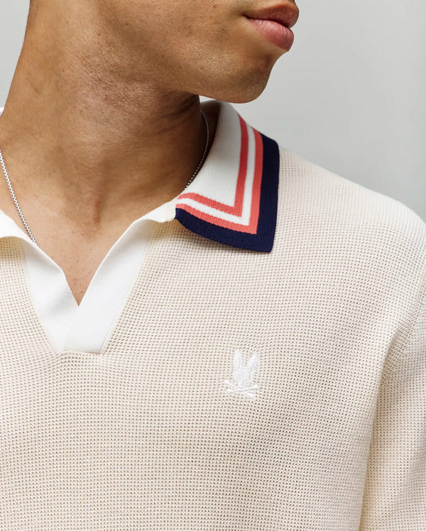 A close-up shows a person wearing the Psycho Bunny MENS CARTER SWEATER POLO (MT0400093), a cream 100% cotton knit Johnny collar shirt with red and navy striped trim, white chest logo embroidery, and a silver necklace. Face partly out of frame.