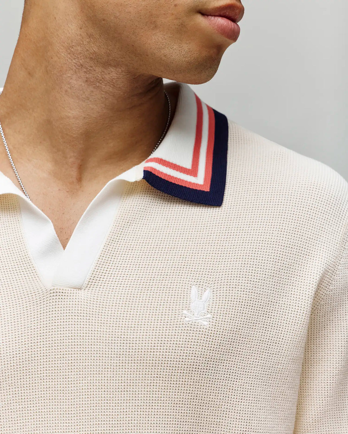A close-up shows a person wearing the Psycho Bunny MENS CARTER SWEATER POLO (MT0400093), a cream 100% cotton knit Johnny collar shirt with red and navy striped trim, white chest logo embroidery, and a silver necklace. Face partly out of frame.