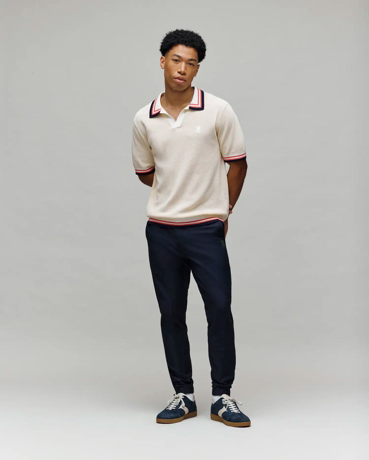 A young man stands against a gray background, wearing the Psycho Bunny MENS CARTER SWEATER POLO - MT0400093 in beige with red and black trim, paired with dark pants and navy sneakers. His hands are behind his back as he faces the camera.