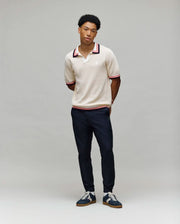 A young man stands against a gray background, wearing the Psycho Bunny MENS CARTER SWEATER POLO - MT0400093 in beige with red and black trim, paired with dark pants and navy sneakers. His hands are behind his back as he faces the camera.