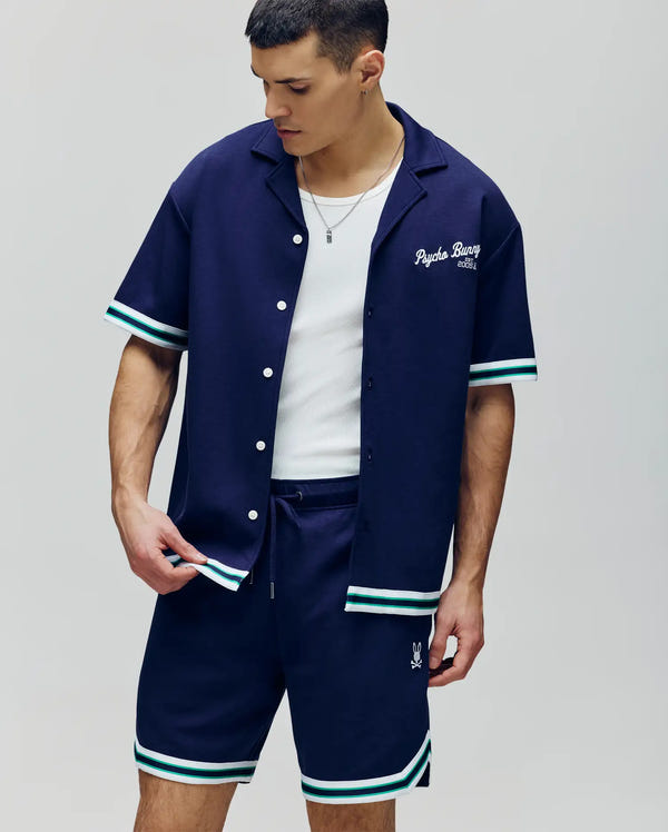 A man wears the Psycho Bunny MENS BASIL TAPED CAMP COLLAR SHIRT (MT0500004) in navy, paired with matching striped shorts, and adjusts his shirt over a white tank top. The set is crafted from a premium cotton-modal stretch blend.