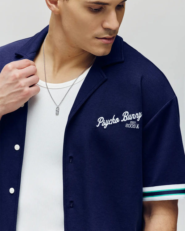 A man wears the Psycho Bunny MENS BASIL TAPED CAMP COLLAR SHIRT (navy, short-sleeve, stretch modal blend with "Psycho Bunny Est. 2005" on the chest) over a white tank. He has a silver necklace and looks down to his left.