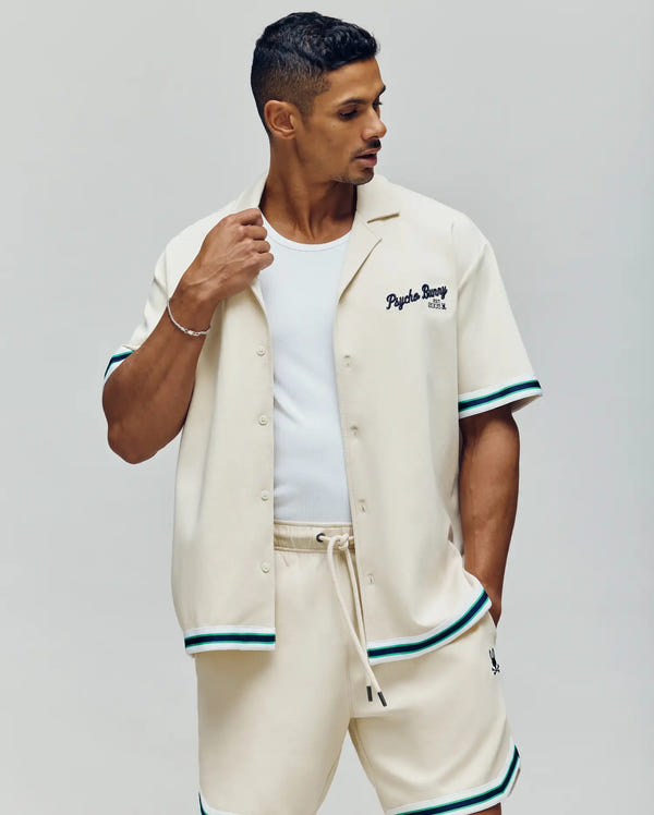 A man with short, dark hair looks to his left, wearing Psycho Bunny’s MENS BASIL TAPED CAMP COLLAR SHIRT (MT0500004), a cream-colored button-up with teal and black striped trim.