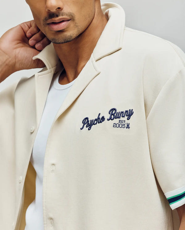 A man wears a cream Psycho Bunny MENS BASIL TAPED CAMP COLLAR SHIRT (MT0500004) with "Psycho Bunny EST. 2005" in blue on the chest over a white tee, raising his left hand behind his neck.