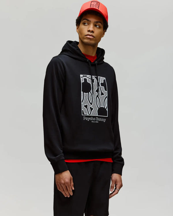 A man wears the Psycho Bunny MENS GLENN HOODIE - MT0500008, featuring a white abstract "Psychic Journey" design, paired with black shorts and a red cap, standing against a plain light gray background.