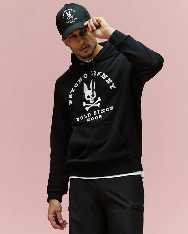 A man in the Psycho Bunny MENS ARCHIVE HOODIE - MT0500009, a black cotton knit pullover with an archive-inspired Bunny and "Psycho Bunny Bold Since 2005" text, plus a matching black cap, stands against a plain pink background.