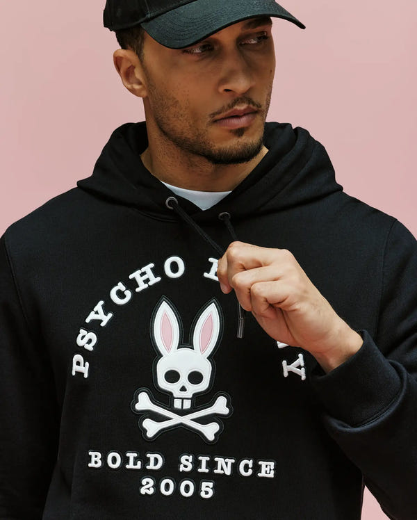 A man in the Psycho Bunny MENS ARCHIVE HOODIE - MT0500009, featuring an Archive-inspired Bunny and "Psycho Bunny Bold Since 2005" text, stands against a pink background adjusting the hoodie string and wearing a black cap.