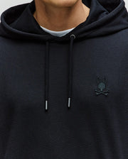 Close-up of a person wearing the Psycho Bunny MENS KAI HOODIE - MT0500019, a black twill-textured hoodie with a small embroidered Bunny logo above crossed bones on the left chest. Only the lower face and torso are visible.