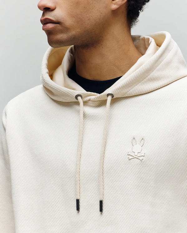 A person wears the cream-colored Psycho Bunny MENS KAI HOODIE - MT0500019, featuring a twill texture, drawstring hood, and embroidered Bunny logo with crossbones on the chest. Their face is partially visible, turned to the side.