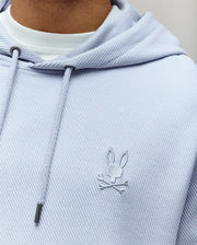 A close-up of the Psycho Bunny MENS KAI HOODIE - MT0500019: a light purple, twill-textured hoodie with a subtle embroidered Bunny logo above crossed bones on the chest, worn over a white tee and finished with metal-tipped drawstrings.