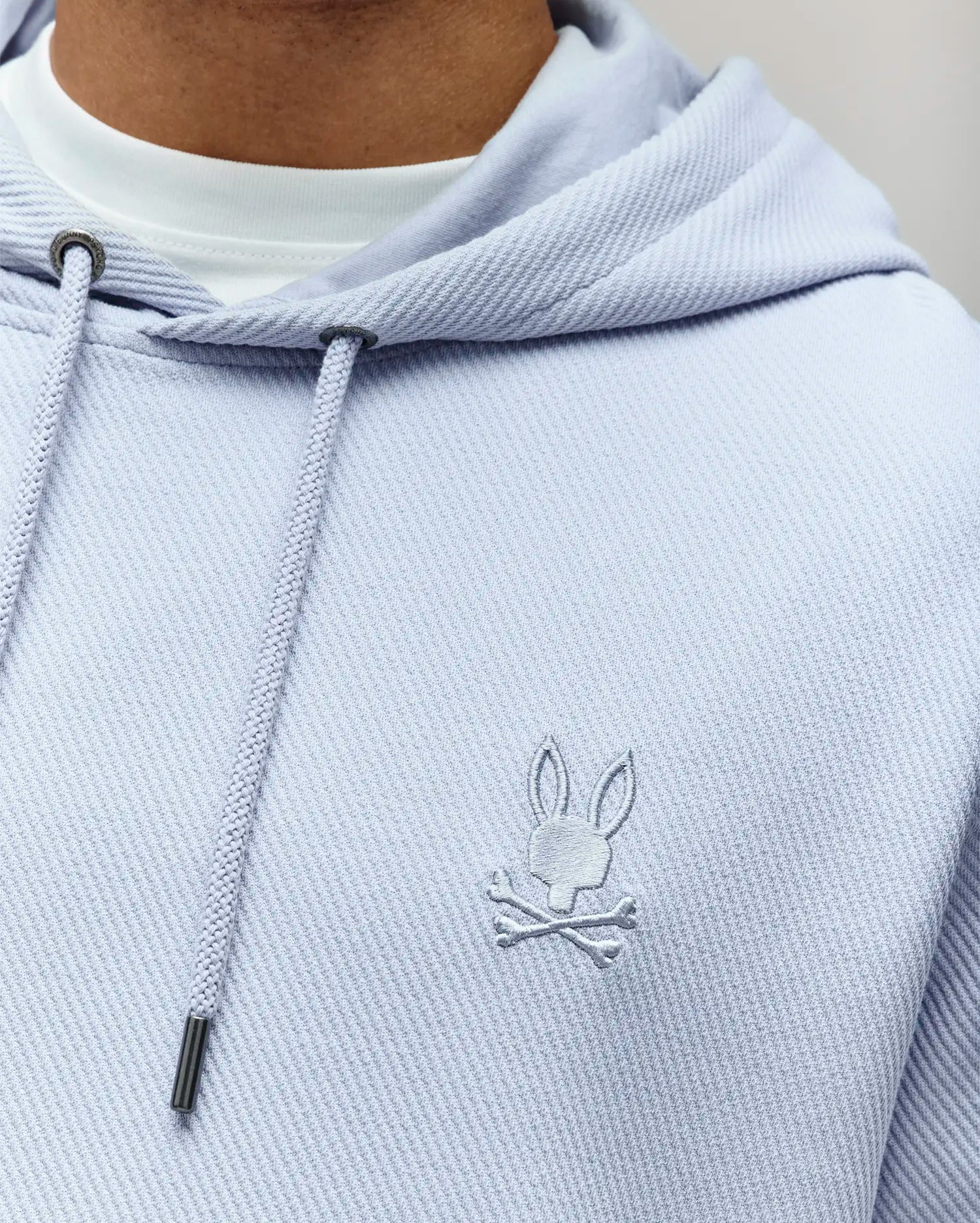 A close-up of the Psycho Bunny MENS KAI HOODIE - MT0500019: a light purple, twill-textured hoodie with a subtle embroidered Bunny logo above crossed bones on the chest, worn over a white tee and finished with metal-tipped drawstrings.
