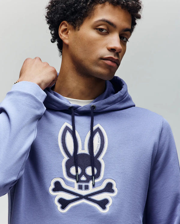 Wearing the Psycho Bunny MENS RYDER BUNNY HOODIE (MT0500029) in light purple, featuring a bold skull-and-crossbones with bunny ears graphic, a confident person poses with one hand near their neck—perfect for stylish lounging.