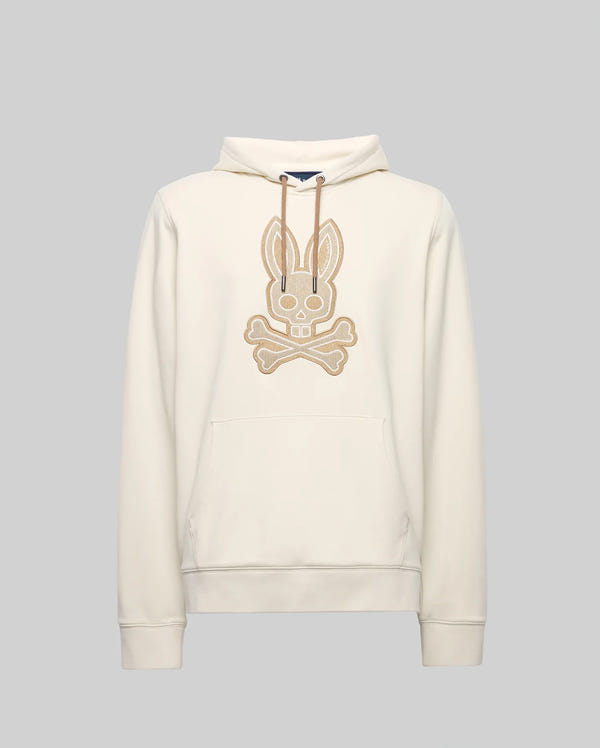 The Psycho Bunny MENS RYDER BUNNY HOODIE (MT0500029) is a cream hoodie with a large beige embroidered Bunny skull-and-crossbones patch, drawstring hood, and kangaroo pocket—ideal for lounging. Shown against a plain gray background.