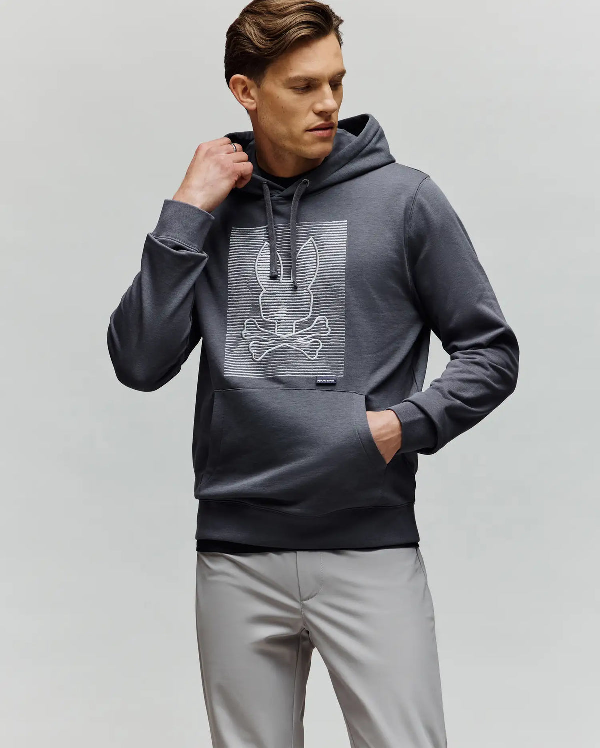 A man wears the Psycho Bunny MENS GRANT HOODIE - MT0500032 in dark gray, made from a premium cotton blend with a bunny and crossbones graphic. He stands against a plain background, one hand in his pocket, the other adjusting his hood.