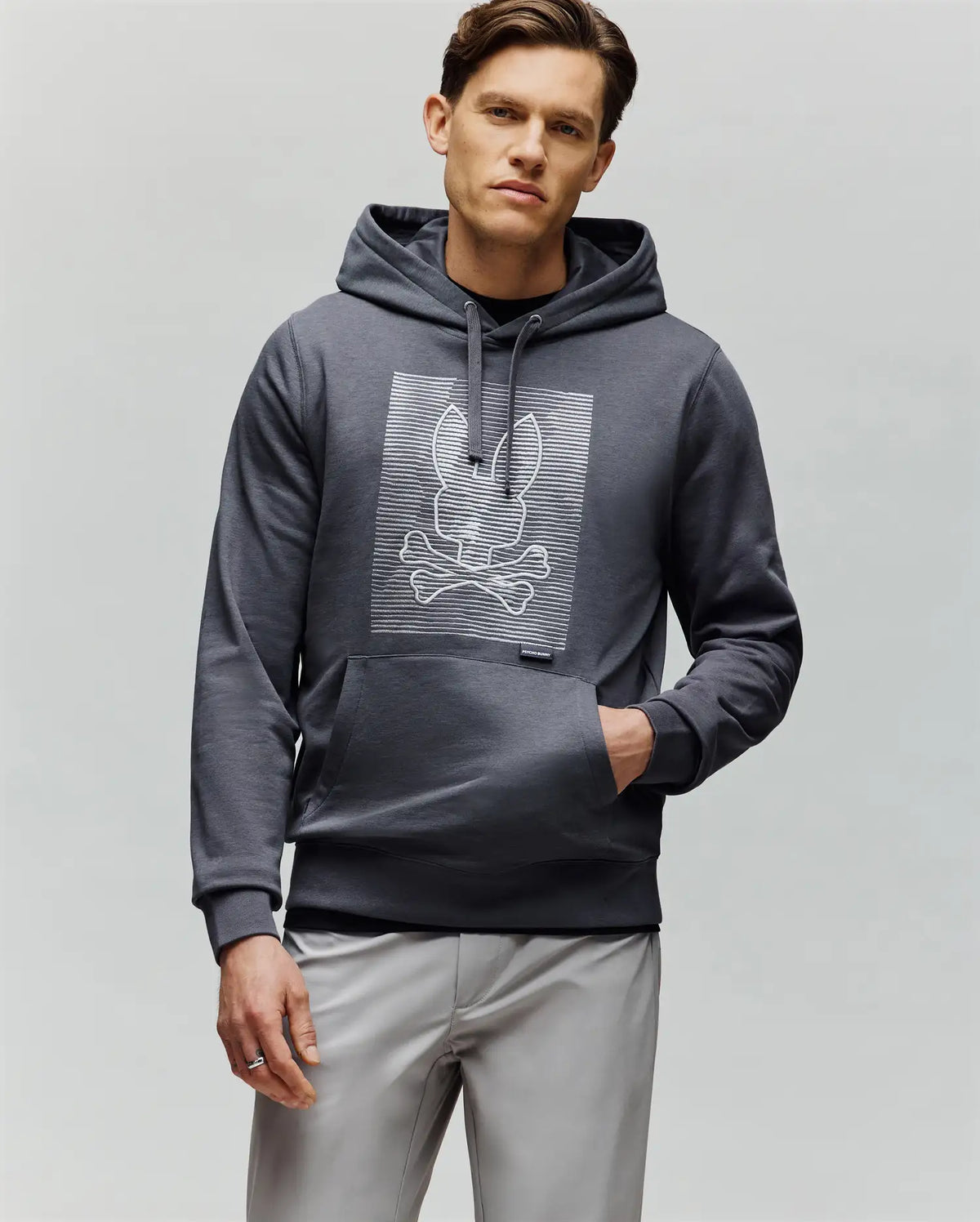 A man wearing the Psycho Bunny MENS GRANT HOODIE - MT0500032, made from a premium cotton blend with a white outlined bunny and crossbones on the chest, stands with one hand in his pocket against a plain light gray background.