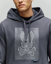 A person wears the Psycho Bunny MENS GRANT HOODIE - MT0500032, a gray premium cotton blend featuring a playful white zig-zag embroidered bunny head above crossed bones. Their face is partially visible.