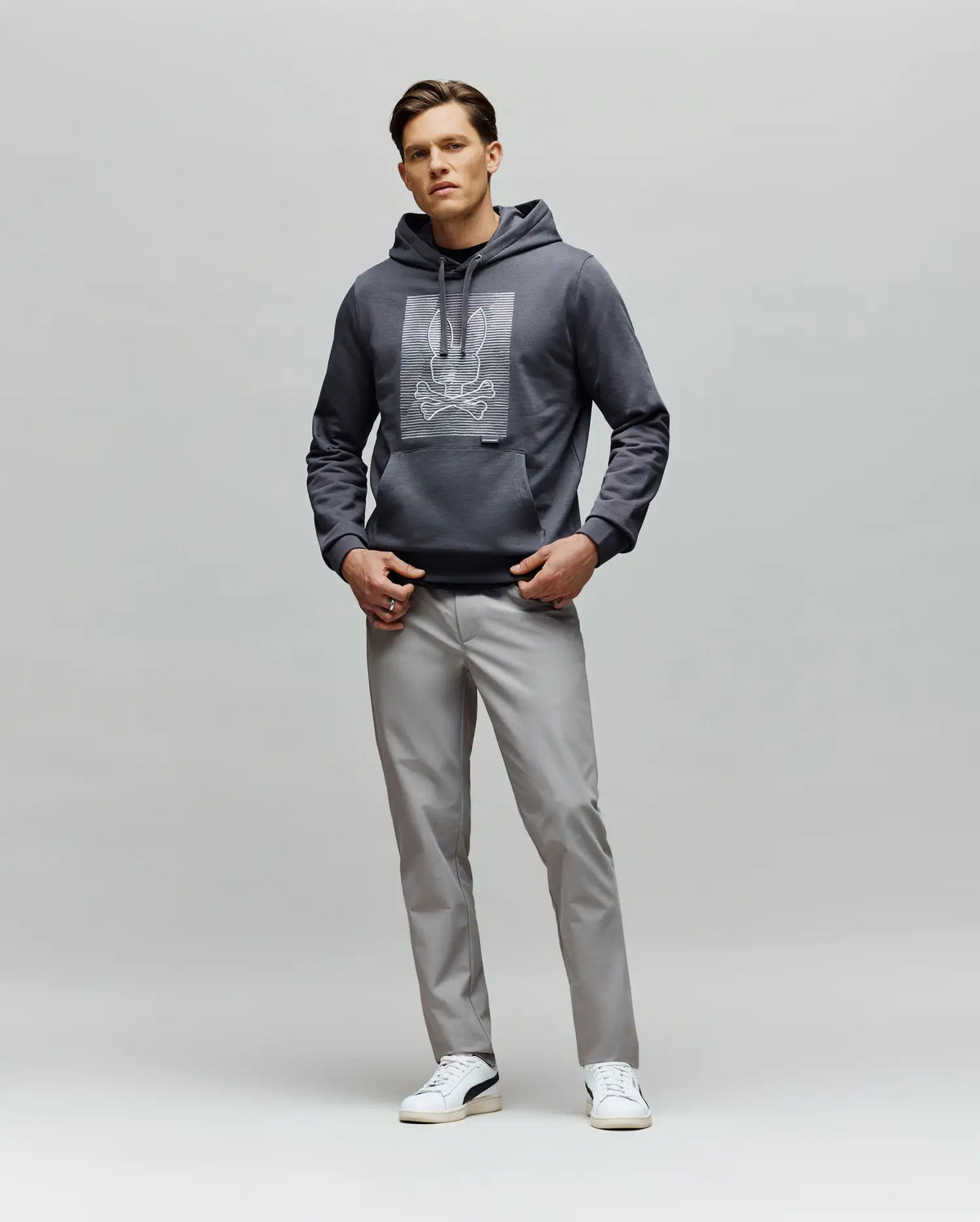 A man faces forward with his hands in his pockets, wearing the Psycho Bunny MENS GRANT HOODIE - MT0500032, light gray pants, and white sneakers against a plain light gray background.