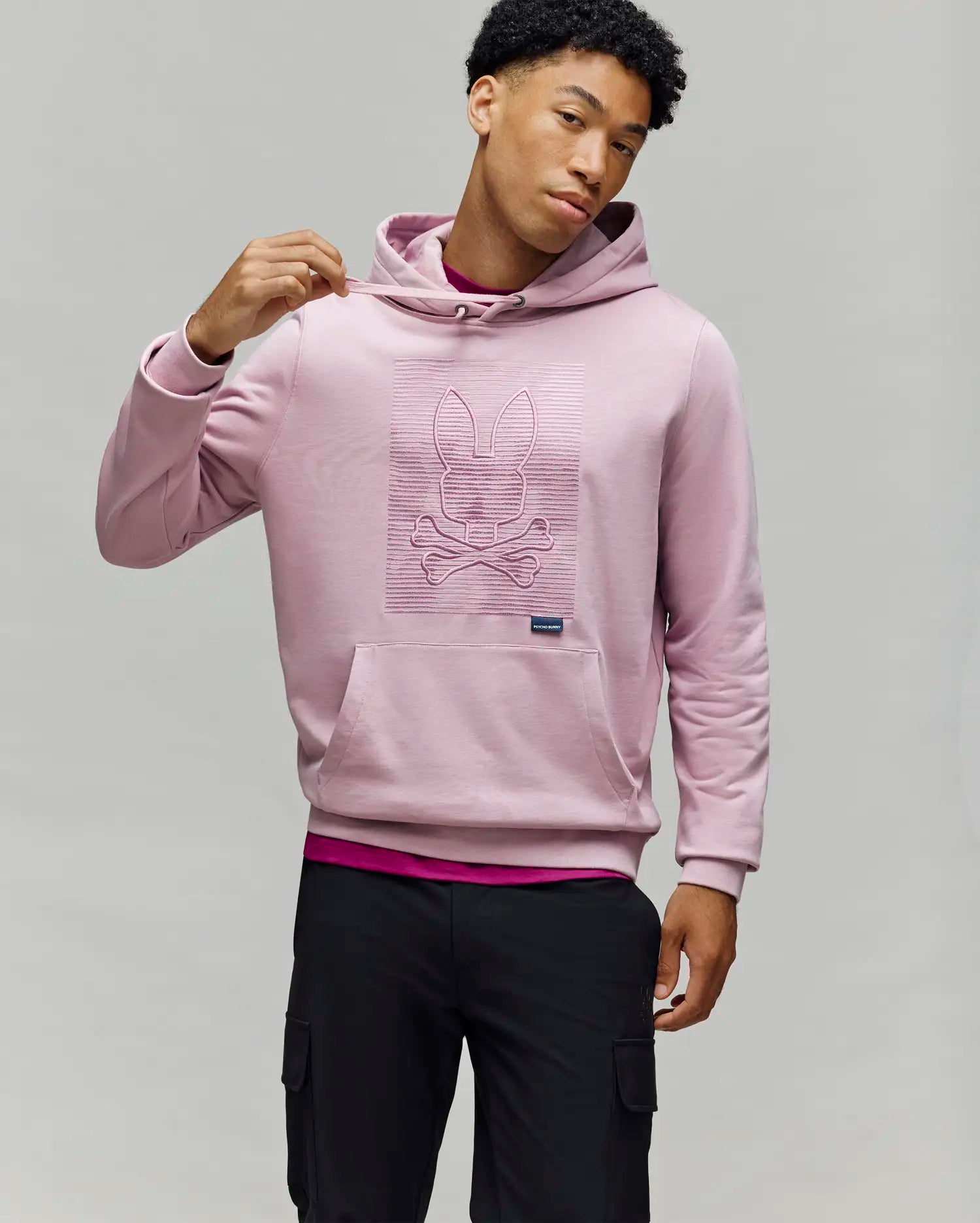 A man in a light pink Psycho Bunny MENS GRANT HOODIE (MT0500032), made from a premium cotton blend with a zig-zag embroidered bunny skull and crossbones, stands against a gray background holding the hood, paired with black cargo pants.