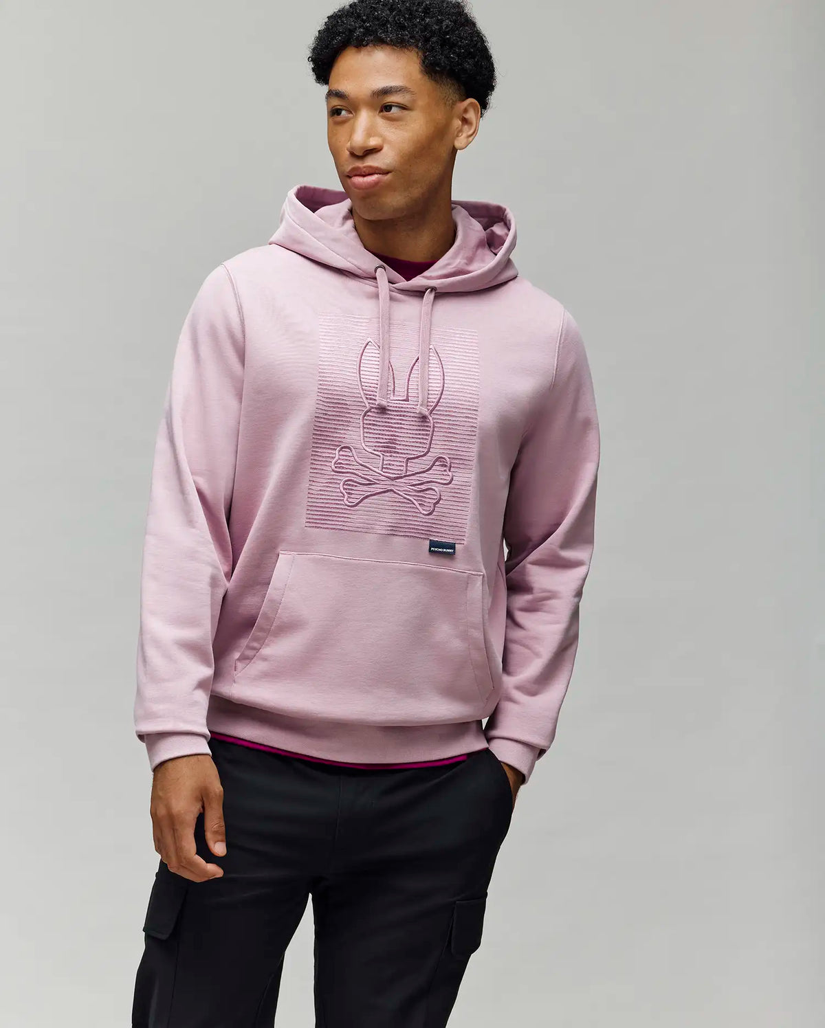A man with short curly hair stands against a gray background, wearing the Psycho Bunny MENS GRANT HOODIE (MT0500032), made from a premium cotton blend and featuring an embossed bunny and crossbones design. One hand is in his pocket as he looks to the side.