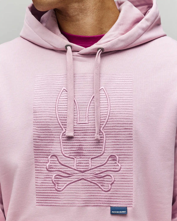A person wears the Psycho Bunny MENS GRANT HOODIE - MT0500032 in light pink, crafted from a premium cotton blend with a raised zig-zag embroidered bunny and crossbones on the chest, hood up, and a small "Psycho Bunny" tag near the hem.