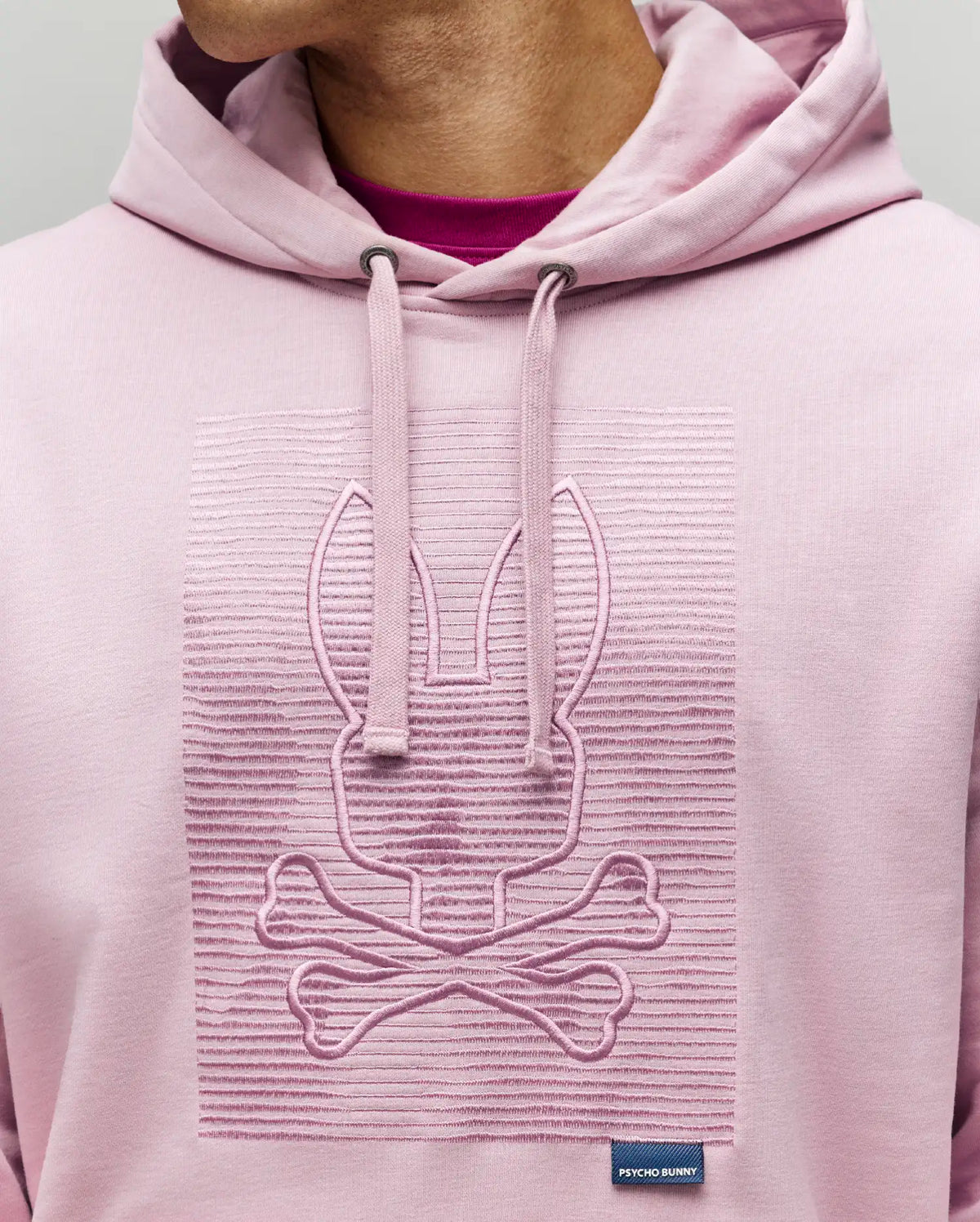 A person wears the Psycho Bunny MENS GRANT HOODIE - MT0500032 in light pink, crafted from a premium cotton blend with a raised zig-zag embroidered bunny and crossbones on the chest, hood up, and a small "Psycho Bunny" tag near the hem.