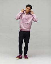 A young man stands against a gray background, wearing the Psycho Bunny MENS GRANT HOODIE - MT0500032 in light pink, paired with black joggers and burgundy sneakers. He holds the hood strings and glances slightly to the side.