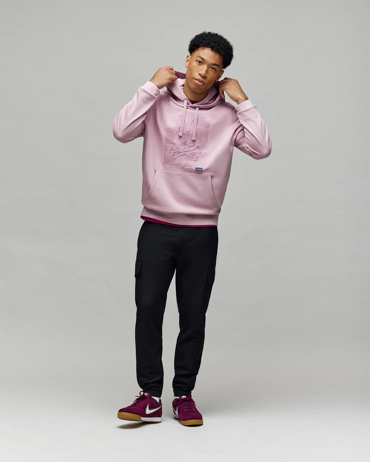 A young man stands against a gray background, wearing the Psycho Bunny MENS GRANT HOODIE - MT0500032 in light pink, paired with black joggers and burgundy sneakers. He holds the hood strings and glances slightly to the side.