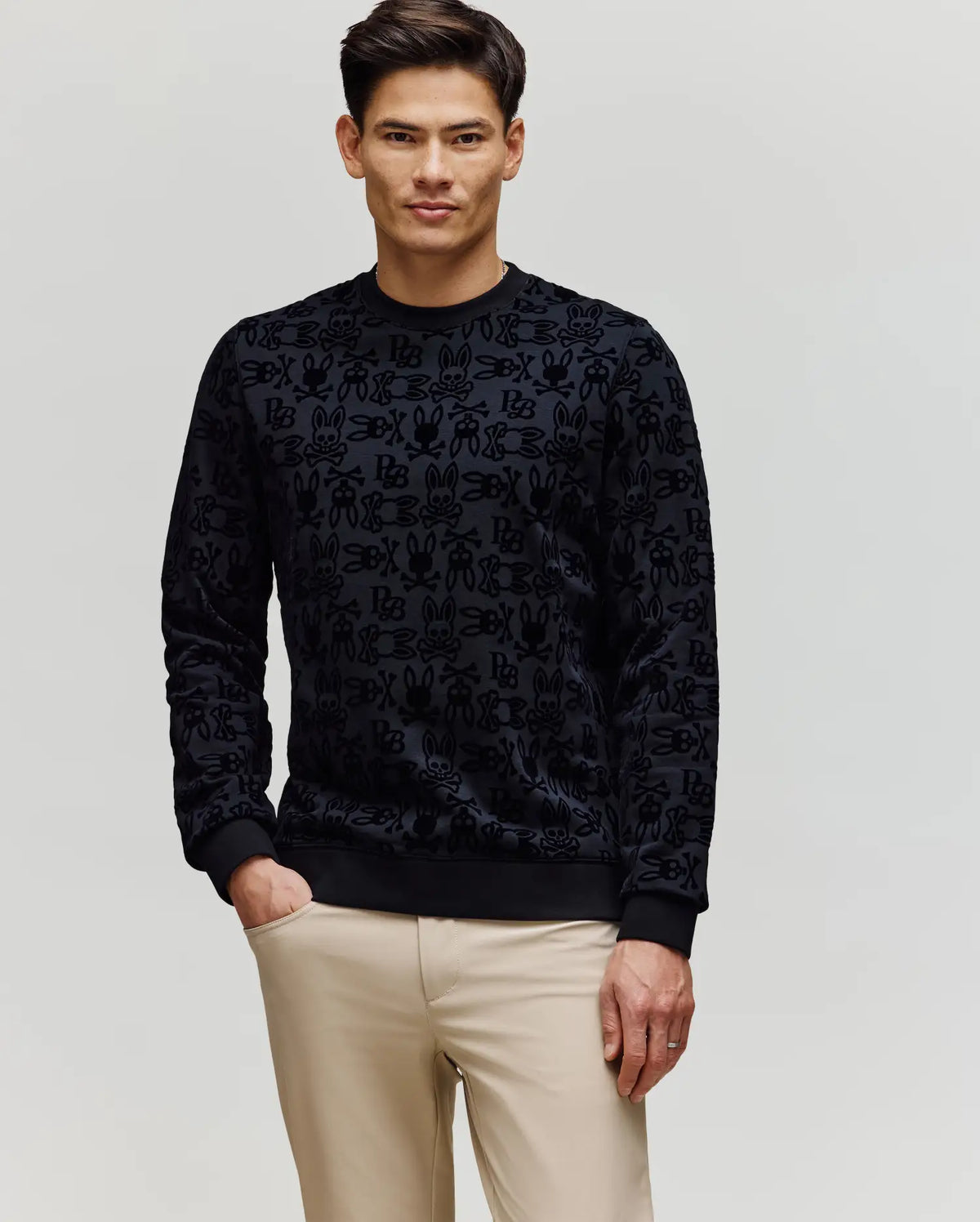 A man with short dark hair wears the Psycho Bunny MENS REED FLOCK CREW (MT0500041) pullover, featuring a subtle Bunny pattern, and light beige pants. He stands against a plain light background with one hand in his pocket.