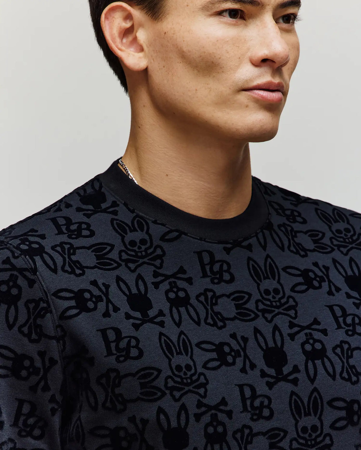 A man with short dark hair models the Psycho Bunny MENS REED FLOCK CREW - MT0500041, a black premium cotton pullover with an allover Bunny and skull-and-crossbones pattern, photographed from the shoulders up against a plain background.