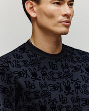 A man with short dark hair models the Psycho Bunny MENS REED FLOCK CREW - MT0500041, a black premium cotton pullover with an allover Bunny and skull-and-crossbones pattern, photographed from the shoulders up against a plain background.