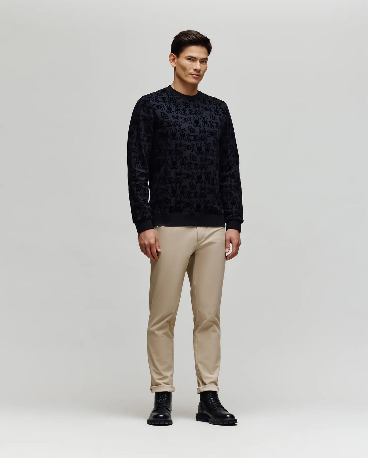 A man stands against a light gray background wearing the Psycho Bunny MENS REED FLOCK CREW (MT0500041), featuring a black premium cotton pullover with an allover Bunny pattern, paired with beige pants and black boots.
