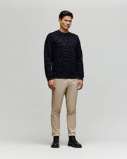 A man stands against a light gray background wearing the Psycho Bunny MENS REED FLOCK CREW (MT0500041), featuring a black premium cotton pullover with an allover Bunny pattern, paired with beige pants and black boots.